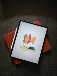 A digital tablet displays an illustration of stylized trees with orange, yellow, and green leaves. The device is placed on a reddish-brown cover, next to a white stylus, resting on a textured dark fabric.