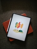A digital tablet displays an illustration of stylized trees with orange, yellow, and green leaves. The device is placed on a reddish-brown cover, next to a white stylus, resting on a textured dark fabric.