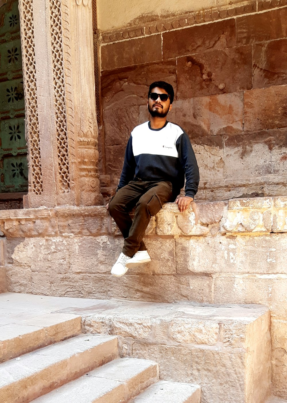 man in black and white long sleeve shirt and brown pants sitting on brown concrete wall