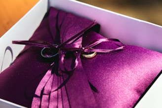 A white box contains a smooth, purple satin pillow adorned with silky purple ribbons. Two gold wedding rings are elegantly placed on the pillow, with the ribbons intertwined around them.