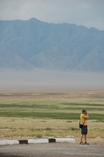 A person stands photographing a vast landscape with rolling plains and distant mountains under an overcast sky. The ground is a mix of green patches and barren earth, while the mountains exhibit a soft blue-gray tone.