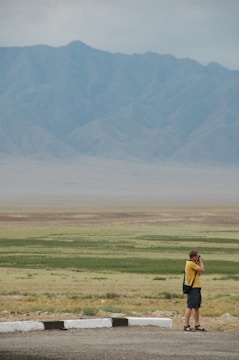 A person stands photographing a vast landscape with rolling plains and distant mountains under an overcast sky. The ground is a mix of green patches and barren earth, while the mountains exhibit a soft blue-gray tone.