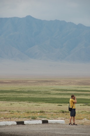 A person stands photographing a vast landscape with rolling plains and distant mountains under an overcast sky. The ground is a mix of green patches and barren earth, while the mountains exhibit a soft blue-gray tone.
