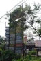 A tall wooden signpost partially covered by foliage displays numerous signs for various hotels and resorts. The signs are colorful, with names like Hotel Hermitage and Jungle Resort, indicating a focus on nature-themed accommodations. The area around the signpost is lush with greenery, and a few buildings and bicycles are visible in the background, suggesting a rural or nature-adjacent setting.