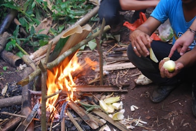 A small campfire surrounded by makeshift supporting sticks is burning. Near the fire, a person is seated, peeling what appears to be a vegetable, possibly bamboo shoots. The ground is covered in leaves and debris, and there is a mix of cut wood and green vegetation around.