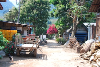 A rural village path lined with tall trees and small wooden structures. A wooden cart is parked on the dirt road, next to stacked firewood. Bright pink flowers and lush greenery create a vibrant, natural setting under a clear sky.