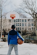 A young person dressed in a blue hoodie holds a basketball while standing on an outdoor court. Leafless trees and a multi-story building can be seen in the background.