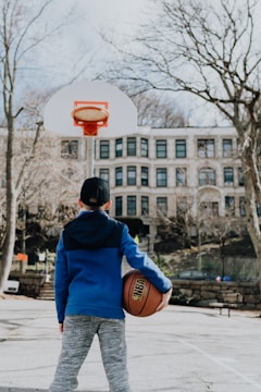 A young person dressed in a blue hoodie holds a basketball while standing on an outdoor court. Leafless trees and a multi-story building can be seen in the background.
