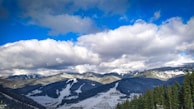 A picturesque mountainous landscape featuring snow-covered slopes and a forested foreground. The sky is partly cloudy with patches of blue peeking through, casting shadows and light across the scene. A winter resort is visible at the base of the slopes.