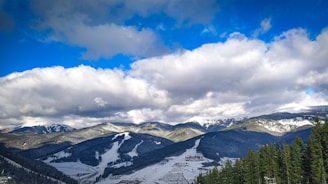 A picturesque mountainous landscape featuring snow-covered slopes and a forested foreground. The sky is partly cloudy with patches of blue peeking through, casting shadows and light across the scene. A winter resort is visible at the base of the slopes.
