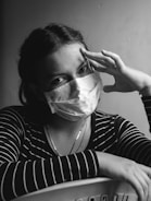 A person wearing a surgical mask looks directly at the camera, with one hand resting on their head and the other arm folded on a surface. The lighting creates a contrast that accentuates the facial features, and the person is dressed in a striped shirt.