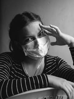 A person wearing a surgical mask looks directly at the camera, with one hand resting on their head and the other arm folded on a surface. The lighting creates a contrast that accentuates the facial features, and the person is dressed in a striped shirt.