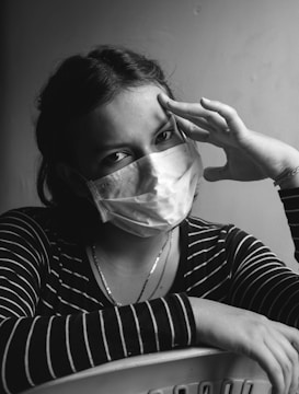 A person wearing a surgical mask looks directly at the camera, with one hand resting on their head and the other arm folded on a surface. The lighting creates a contrast that accentuates the facial features, and the person is dressed in a striped shirt.