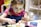 A young child with painted blue designs on their face is focused on painting a small ceramic object, possibly an animal figurine. The child wears a pink outfit with a blue apron and has pigtails. There are various containers of paint in front of them on the table.