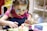 A young child with painted blue designs on their face is focused on painting a small ceramic object, possibly an animal figurine. The child wears a pink outfit with a blue apron and has pigtails. There are various containers of paint in front of them on the table.