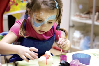 A young child with painted blue designs on their face is focused on painting a small ceramic object, possibly an animal figurine. The child wears a pink outfit with a blue apron and has pigtails. There are various containers of paint in front of them on the table.