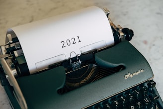 A vintage black typewriter with a sheet of paper inserted, displaying the year 2021 in bold black letters. The typewriter is set on a light-colored, textured surface.