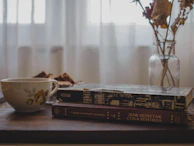 A cozy corner with vintage books and a teacup, a woman’s silhouette in a flowing dress blurred in the background.
