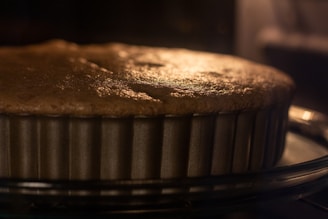 A close-up view of a cake or pie baking in a metal, fluted pan. The surface appears golden brown, indicating it is almost fully baked. The image captures the warm glow, likely from an oven light, creating a cozy and inviting atmosphere.