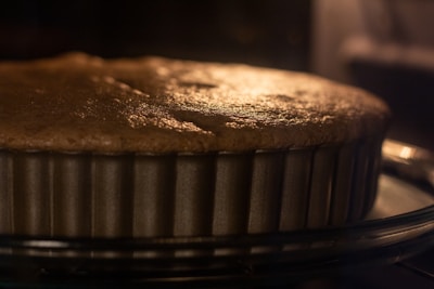 A close-up view of a cake or pie baking in a metal, fluted pan. The surface appears golden brown, indicating it is almost fully baked. The image captures the warm glow, likely from an oven light, creating a cozy and inviting atmosphere.
