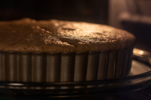 A close-up view of a cake or pie baking in a metal, fluted pan. The surface appears golden brown, indicating it is almost fully baked. The image captures the warm glow, likely from an oven light, creating a cozy and inviting atmosphere.