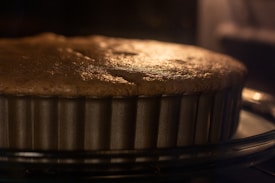 A close-up view of a cake or pie baking in a metal, fluted pan. The surface appears golden brown, indicating it is almost fully baked. The image captures the warm glow, likely from an oven light, creating a cozy and inviting atmosphere.