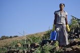 A person wearing a headscarf and gloves stands on a terraced hillside garden holding a blue watering can. The garden is filled with various plants, and the background shows a clear blue sky and a rural landscape.