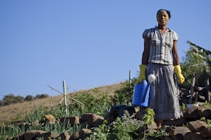 A person wearing a headscarf and gloves stands on a terraced hillside garden holding a blue watering can. The garden is filled with various plants, and the background shows a clear blue sky and a rural landscape.
