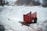 A red wagon with a small loader is positioned in a snow-covered yard. The surroundings include a chain-link fence, trees, and a distant house, suggesting a residential area. The scene captures a winter landscape with visible dirt patches amidst the snow.