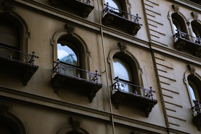 A detailed view of a building's facade featuring multiple arched windows with intricately designed iron railings. The architecture appears historic, with stone or concrete material making up the structure. Reflections of trees are visible in some of the windowpanes, adding a touch of nature to the urban setting.
