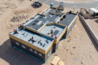 Aerial view of a construction site featuring a large building with workers on the flat roof installing materials. The building is surrounded by sand and construction debris. Nearby, there is a pile of rocks and a small truck. The area is mostly barren with a road curving in the background.