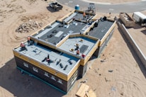 Aerial view of a construction site featuring a large building with workers on the flat roof installing materials. The building is surrounded by sand and construction debris. Nearby, there is a pile of rocks and a small truck. The area is mostly barren with a road curving in the background.
