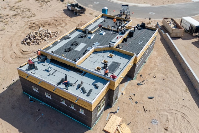 Aerial view of a construction site featuring a large building with workers on the flat roof installing materials. The building is surrounded by sand and construction debris. Nearby, there is a pile of rocks and a small truck. The area is mostly barren with a road curving in the background.