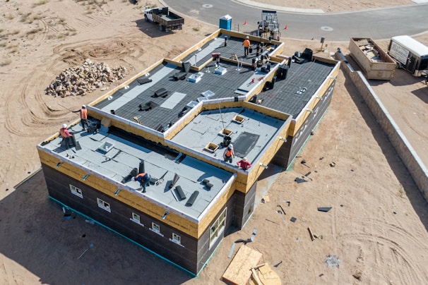 Aerial view of a construction site featuring a large building with workers on the flat roof installing materials. The building is surrounded by sand and construction debris. Nearby, there is a pile of rocks and a small truck. The area is mostly barren with a road curving in the background.