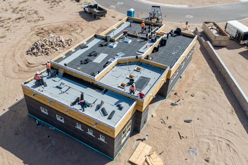 Aerial view of a construction site featuring a large building with workers on the flat roof installing materials. The building is surrounded by sand and construction debris. Nearby, there is a pile of rocks and a small truck. The area is mostly barren with a road curving in the background.