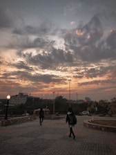 A paved walkway bordered by stone walls extends into the distance, under a dramatic sky filled with dark, swirling clouds tinged with orange hues from the setting sun. Two people, one in the foreground, walk toward a well-lit area. Tall streetlights and a building sign reading 'Kidney' are visible in the background.