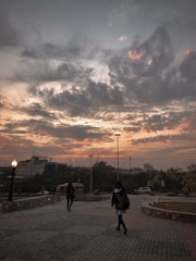 A paved walkway bordered by stone walls extends into the distance, under a dramatic sky filled with dark, swirling clouds tinged with orange hues from the setting sun. Two people, one in the foreground, walk toward a well-lit area. Tall streetlights and a building sign reading 'Kidney' are visible in the background.