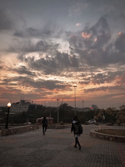 A paved walkway bordered by stone walls extends into the distance, under a dramatic sky filled with dark, swirling clouds tinged with orange hues from the setting sun. Two people, one in the foreground, walk toward a well-lit area. Tall streetlights and a building sign reading 'Kidney' are visible in the background.