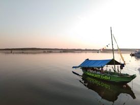 A small boat with a blue tarp canopy is moored in calm waters. The boat has colorful flags on a mast and the word 'Wakatobi' visible on its side. The horizon shows a flat landscape under a clear sky during sunset or sunrise, casting a warm reflection on the water. Other boats can be seen faintly in the background, suggesting a peaceful waterfront scene.