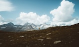 A solitary hiker wearing a bright yellow jacket stands on a rocky hillside. In the background, dramatic alpine mountains rise under a vast sky filled with large, white fluffy clouds. The landscape conveys a sense of expansive wilderness and adventure.