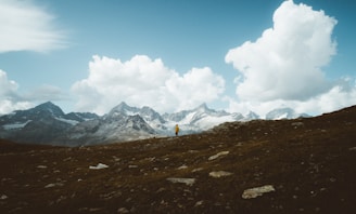 A solitary hiker wearing a bright yellow jacket stands on a rocky hillside. In the background, dramatic alpine mountains rise under a vast sky filled with large, white fluffy clouds. The landscape conveys a sense of expansive wilderness and adventure.
