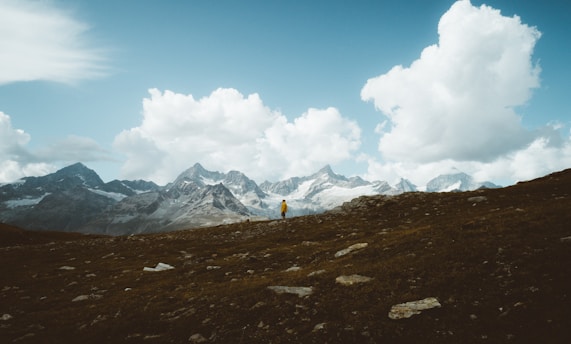 A solitary hiker wearing a bright yellow jacket stands on a rocky hillside. In the background, dramatic alpine mountains rise under a vast sky filled with large, white fluffy clouds. The landscape conveys a sense of expansive wilderness and adventure.