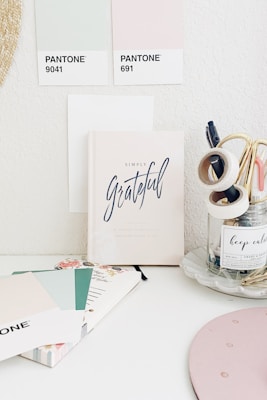 A minimalistic workspace featuring pastel-colored Pantone swatches and a notebook titled 'Simply Grateful.' The desk also has stationery items such as tapes and pens organized in a clear jar and a notebook with floral designs. The space gives off a calm and organized vibe.
