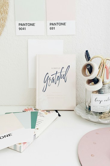 A minimalistic workspace featuring pastel-colored Pantone swatches and a notebook titled 'Simply Grateful.' The desk also has stationery items such as tapes and pens organized in a clear jar and a notebook with floral designs. The space gives off a calm and organized vibe.
