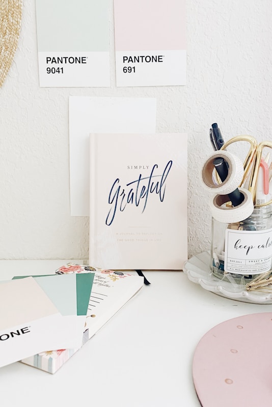 A minimalistic workspace featuring pastel-colored Pantone swatches and a notebook titled 'Simply Grateful.' The desk also has stationery items such as tapes and pens organized in a clear jar and a notebook with floral designs. The space gives off a calm and organized vibe.