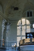 An ornate interior features detailed sculptures of figures integrated into the walls, bathed in soft natural light. Elaborate carvings and decorative moldings adorn the space, highlighted by sunlight casting shadows. A window is partially visible above a richly decorated balcony railing.