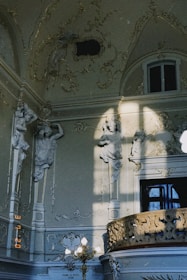 An ornate interior features detailed sculptures of figures integrated into the walls, bathed in soft natural light. Elaborate carvings and decorative moldings adorn the space, highlighted by sunlight casting shadows. A window is partially visible above a richly decorated balcony railing.