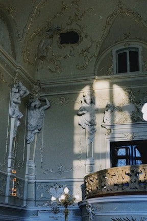 An ornate interior features detailed sculptures of figures integrated into the walls, bathed in soft natural light. Elaborate carvings and decorative moldings adorn the space, highlighted by sunlight casting shadows. A window is partially visible above a richly decorated balcony railing.