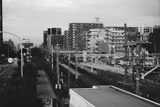 A black and white urban scene featuring a railway track running through a cityscape. High-rise residential buildings dominate the background, while an electric railway infrastructure is visible throughout the scene. Road signs, lampposts, and overhead electrical wires add complexity to the frame.