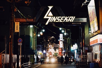 A bustling street scene in a Japanese city at night with neon lights illuminating the area. The Sinsekai sign is prominently displayed, surrounded by various shops and advertisements. Several people are walking along the sidewalk, and a few vehicles are visible.